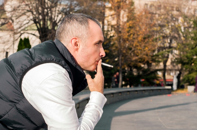 Photo beau jeune homme élégant qui fume à l'extérieur en milieu urbain, en détournant les yeux. bel homme blanc fumant une sigarette en plein air en journée ensoleillée