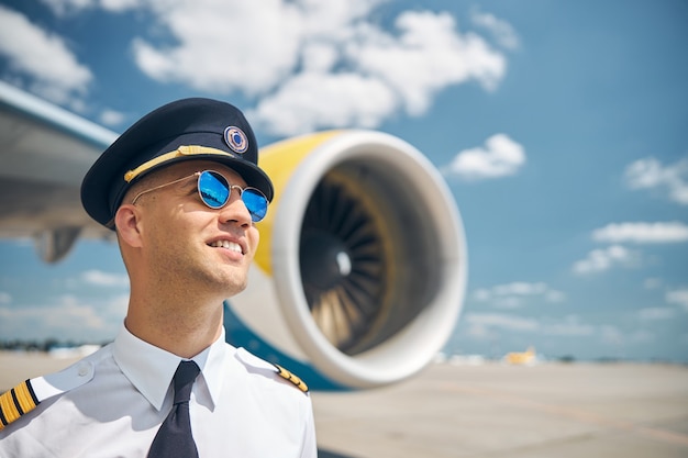 Beau jeune homme employé de compagnie aérienne regardant loin et souriant tout en se tenant à l'aéroport avec avion et ciel sur fond flou