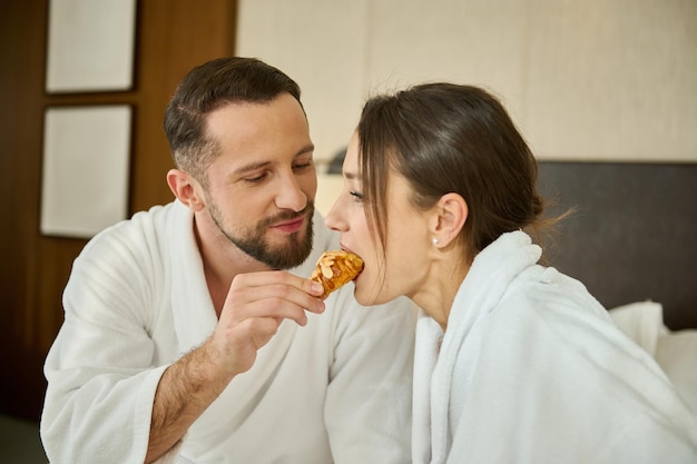 Un beau jeune homme européen séduisant en peignoir nourrit sa femme bien-aimée, partenaire de délicieuses pâtisseries cachées tout en prenant son petit-déjeuner au lit. Romance, amour, attirance. Petit déjeuner au lit pour deux.