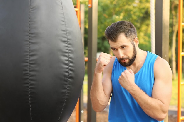 Beau jeune homme faisant de l'exercice avec un sac de boxe dans le parc