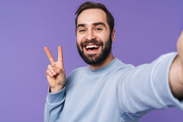 un beau jeune homme heureux et positif isolé sur un mur violet prend un selfie par caméra montrant la paix.