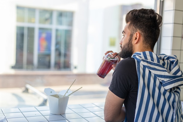 Beau jeune homme hipster buvant un cocktail dans un café et regardant par la fenêtre.