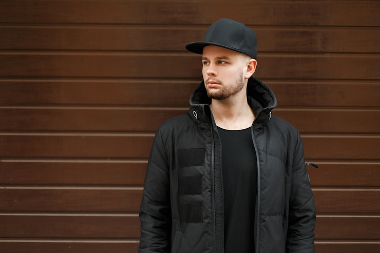 Photo beau jeune homme hipster dans une casquette de baseball élégante noire et veste d'hiver se tient près du mur