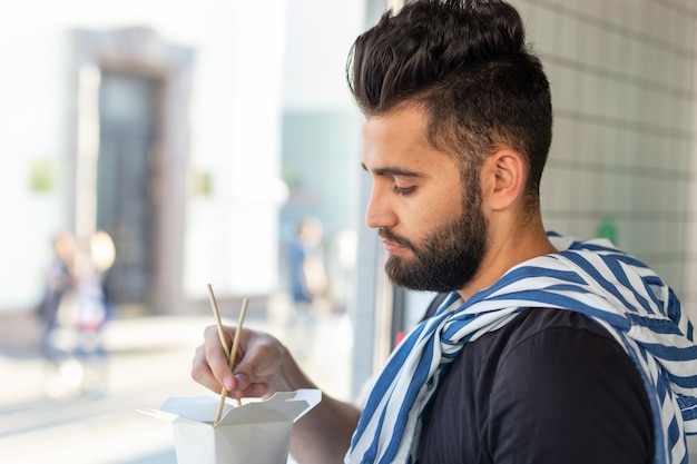 Beau jeune homme hipster mangeant des nouilles chinoises à l'aide de baguettes en bois assis dans un café et regardant dans la fenêtre. Concept de cuisine asiatique.