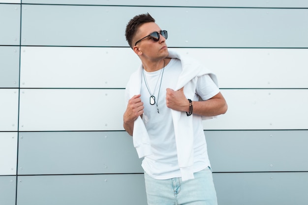 Beau jeune homme hipster avec un sweat-shirt blanc dans un T-shirt vintage dans des lunettes de soleil élégantes avec une coiffure à la mode posant