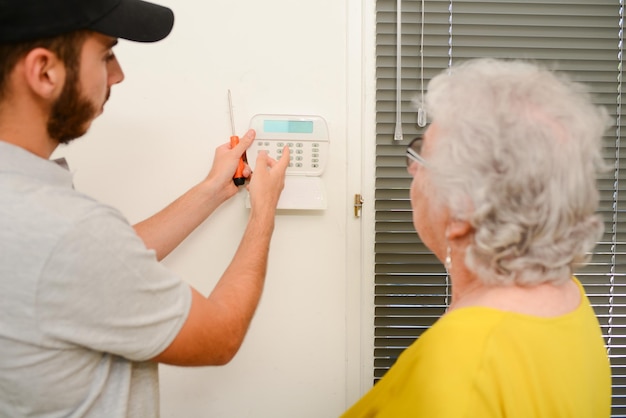 Beau jeune homme installant une caméra anti-cambriolage et une sirène d'alarme de sécurité dans une maison de femme âgée