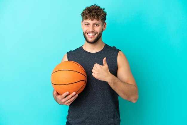 Beau jeune homme jouant au basket-ball isolé sur fond bleu donnant un coup de pouce geste