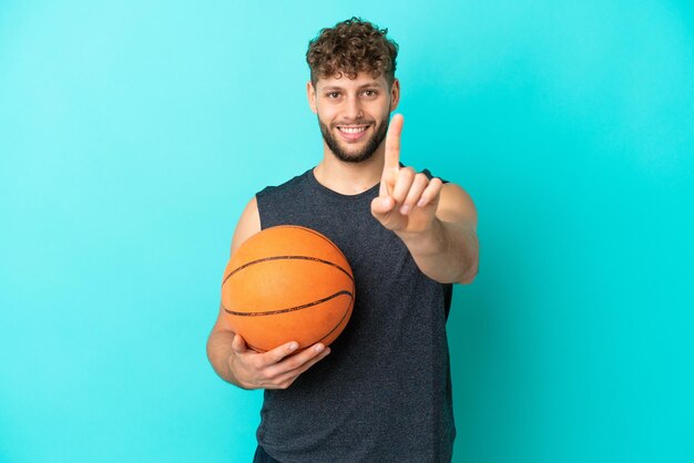 Beau jeune homme jouant au basket-ball isolé sur fond bleu montrant et levant un doigt