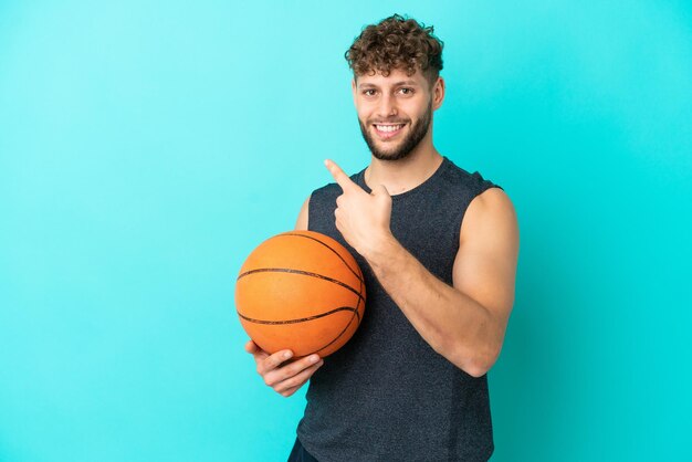 Beau jeune homme jouant au basket-ball isolé sur fond bleu pointant vers l'arrière