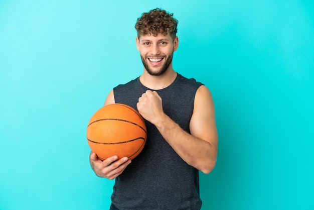 Beau jeune homme jouant au basketball isolé sur fond bleu célébrant une victoire