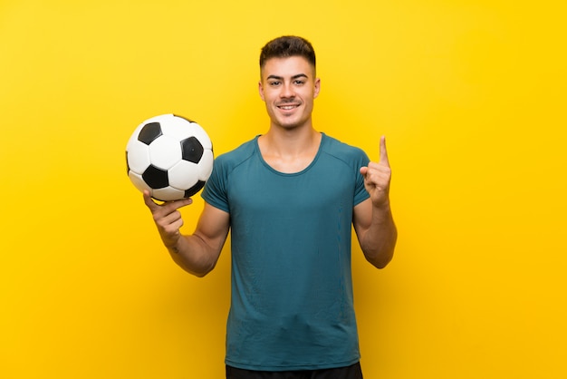 Beau jeune homme joueur de football sur un mur jaune isolé pointant vers le haut une excellente idée