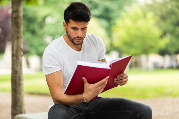 Beau jeune homme lisant un livre sur un banc dans le parc