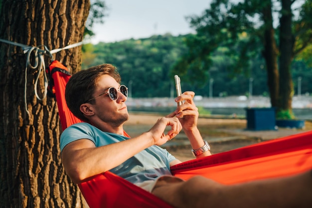 Beau jeune homme à lunettes de soleil couché au coucher du soleil dans un hamac et à l'aide d'un smartphone