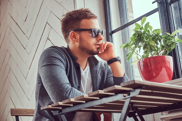 Un beau jeune homme en lunettes de soleil et vêtements décontractés assis dans un café.