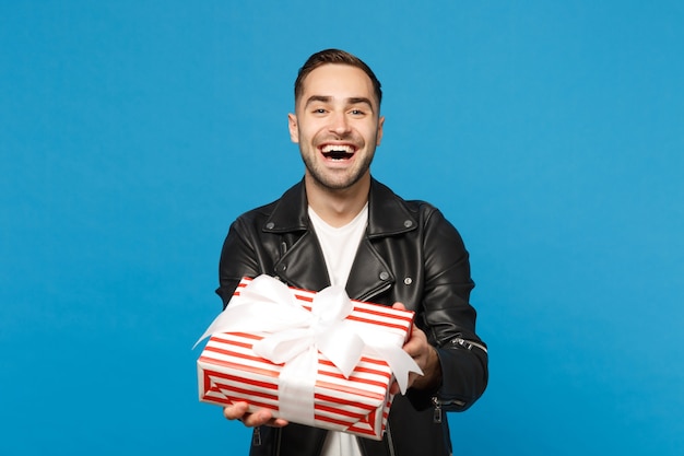 Beau jeune homme mal rasé élégant en blouson de cuir noir t-shirt blanc tenir une boîte-cadeau isolée sur fond de mur bleu portrait en studio. Concept de mode de vie des émotions sincères des gens. Maquette de l'espace de copie.