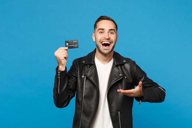 Beau jeune homme mal rasé élégant en blouson de cuir noir t-shirt blanc tenir en main une carte bancaire de crédit isolée sur fond de mur bleu portrait en studio. Concept de mode de vie des gens. Maquette de l'espace de copie.