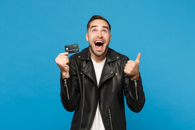 Beau jeune homme mal rasé élégant en blouson de cuir noir t-shirt blanc tenir en main une carte bancaire de crédit isolée sur fond de mur bleu portrait en studio. Concept de mode de vie des gens. Maquette de l'espace de copie.