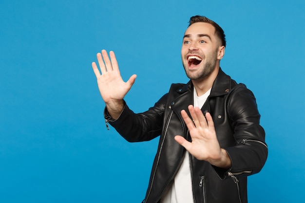 Beau jeune homme mal rasé élégant en t-shirt blanc avec veste en cuir noir regardant de côté isolé sur fond de mur bleu portrait en studio. Concept de mode de vie des émotions sincères des gens. Maquette de l'espace de copie.