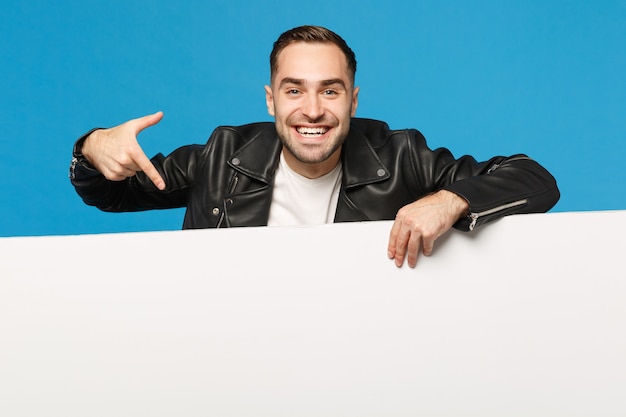 Un beau jeune homme mal rasé tient un grand panneau d'affichage blanc vide pour le contenu promotionnel isolé sur un portrait de studio de fond de mur bleu. Concept de mode de vie des émotions sincères des gens. Maquette de l'espace de copie