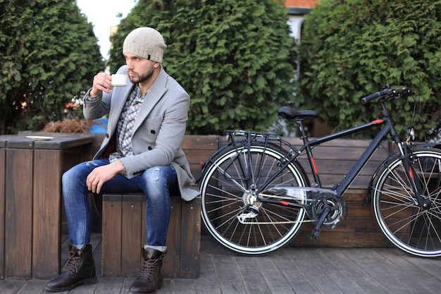 Beau jeune homme en manteau gris et chapeau assis sur un banc détendu en buvant du café et en pensant près de son vélo.