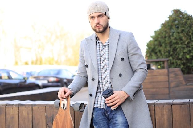 Beau jeune homme en manteau gris et chapeau buvant du café, se reposant, debout avec un longboard. Concept de skateboard urbain.