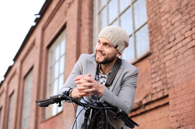 Beau jeune homme en manteau gris et chapeau debout avec son vélo.