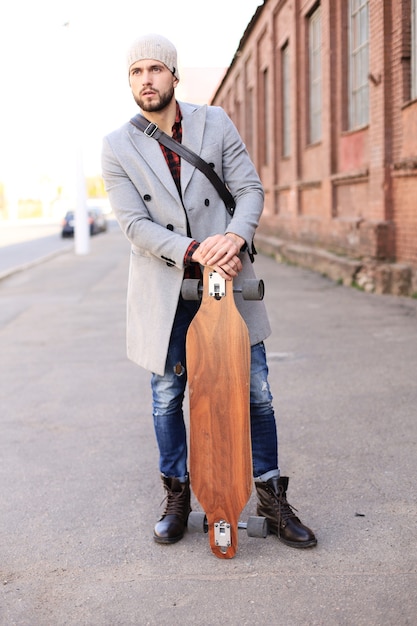 Beau jeune homme en manteau gris et chapeau marchant dans la rue, à l'aide de longboard.