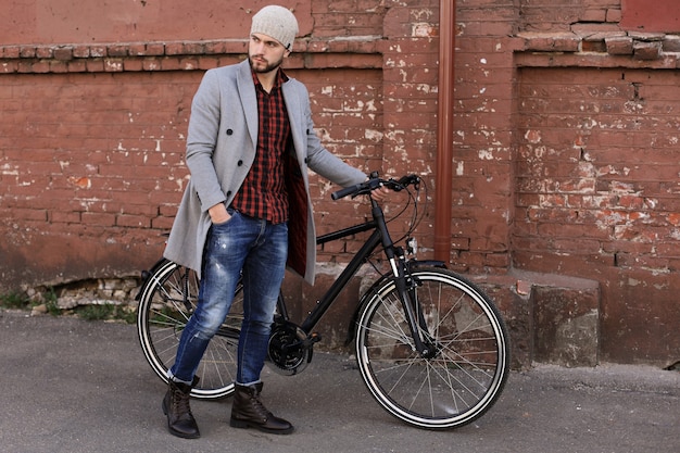 Beau jeune homme en manteau gris et chapeau poussant son vélo dans une rue de la ville.