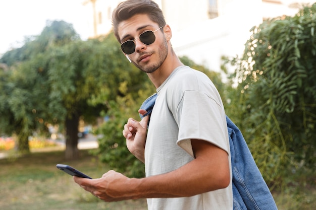 Beau jeune homme marchant à l'extérieur en écoutant de la musique dans la rue à l'aide de téléphone portable et de bavarder