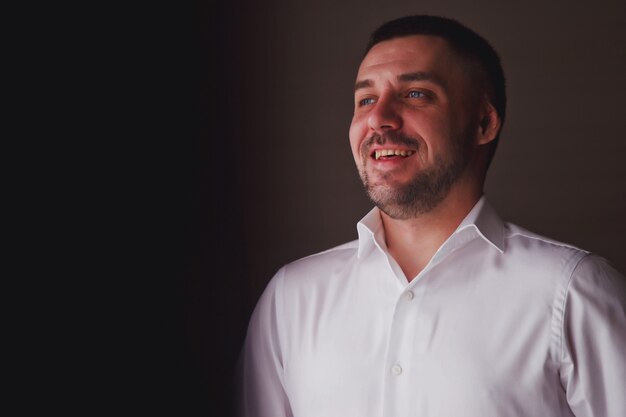 Beau jeune homme marié habille une chemise blanche à la fenêtre de la chambre d'hôtel, sourit avec un regard sûr lui-même. Un mec mignon se prépare à rencontrer la mariée. Concept de bonheur et de luxe marié. Espace de copie