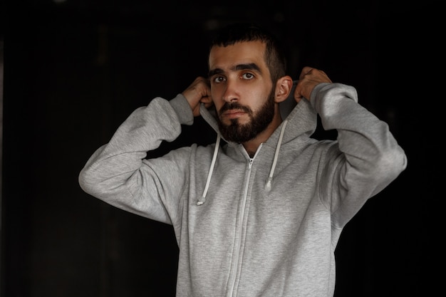 Beau jeune homme de mode avec barbe en sweat à capuche gris contre un mur sombre