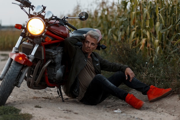 Beau jeune homme à la mode dans une veste militaire à la mode avec des baskets se trouve près d'une moto avec de la lumière dans un champ de maïs dans la soirée