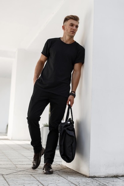 Beau jeune homme à la mode dans des vêtements élégants noirs avec un sac noir posant près du mur blanc