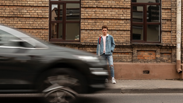 Beau jeune homme moderne en veste en jean bleu élégant se tient dans la rue près de la route devant les voitures en mouvement. Un mec à la mode dans des vêtements de jeans décontractés pour jeunes à la mode marche à l'extérieur. Le trafic de la ville.