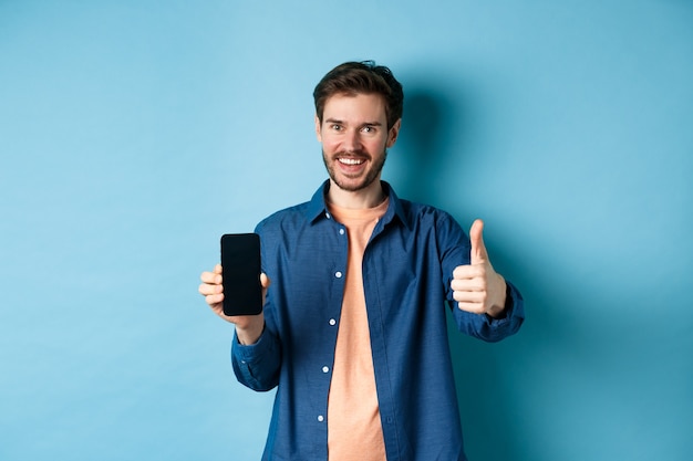 Beau jeune homme montrant le geste du pouce vers le haut et l'écran du smartphone vide, recommandant une application ou une entreprise, debout sur fond bleu.