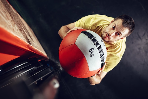 Beau jeune homme musclé faisant de l'exercice avec un ballon mural au gymnase. Vue de dessus. Regarder la caméra.