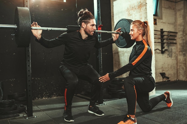 Beau jeune homme musclé fait des exercices de squat avec des haltères aidés par un entraîneur personnel au gymnase.