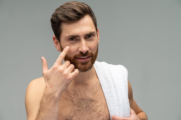 Beau jeune homme nu barbu avec une peau parfaite appliquant une crème hydratante pour le visage isolé sur fond gris portrait en studio Soins de la peau concept de procédures cosmétiques de soins de santé