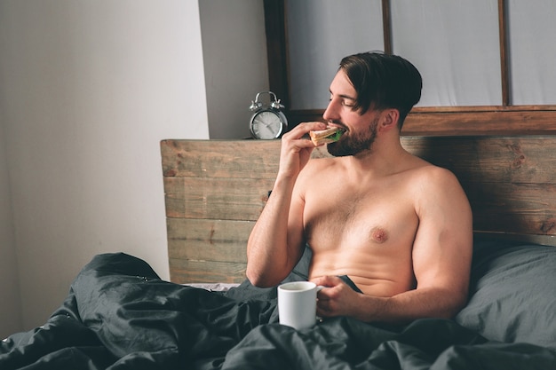 Beau jeune homme nu souriant tenant une tasse de café et regardant ailleurs alors qu'il était assis sur le lit.