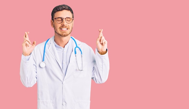 Beau jeune homme avec ours portant l'uniforme de médecin gesticulant doigt croisé souriant avec espoir et les yeux fermés. chance et concept superstitieux.