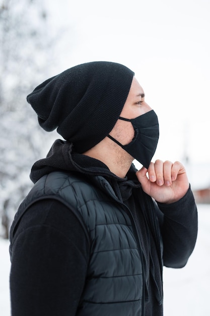 Beau jeune homme portant un masque noir protecteur à l'extérieur sur fond blanc. Mec à la mode dans une tenue élégante noire avec un gilet, un sweat à capuche et un chapeau un jour d'hiver enneigé. Façon COVID-19