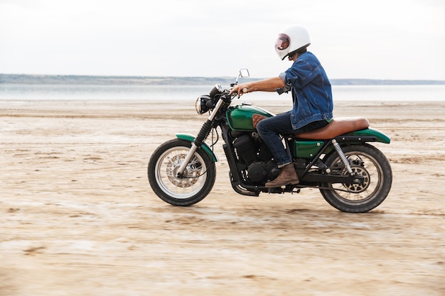Beau jeune homme portant une tenue décontractée faisant de la moto à la plage, portant un casque