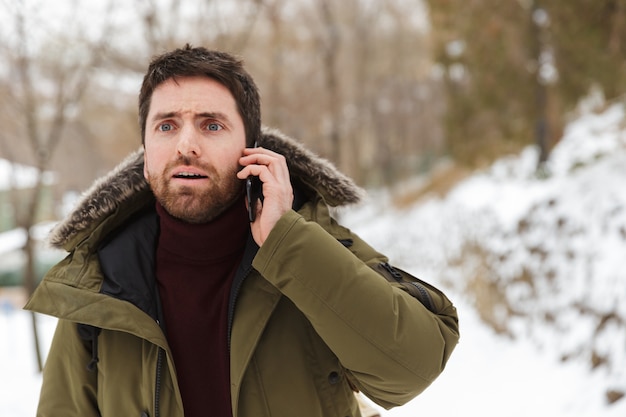 Beau jeune homme portant une veste d'hiver debout à l'extérieur, parler au téléphone mobile