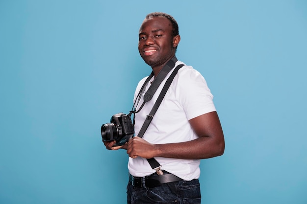 Beau jeune homme portant des vêtements de mode tout en ayant un appareil photo. Photographe confiant ayant un appareil photo professionnel souriant chaleureusement tout en se tenant sur fond bleu.