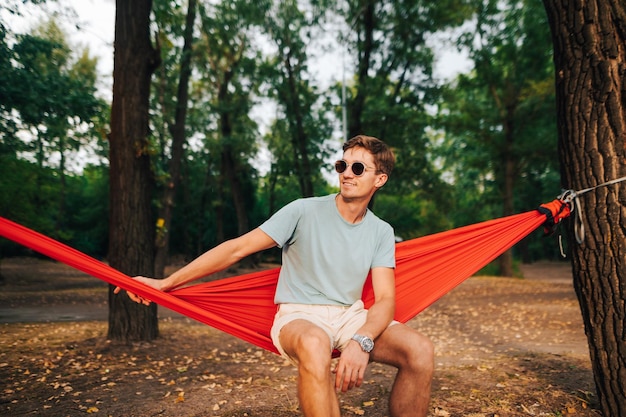 Beau jeune homme positif en lunettes de soleil et vêtements élégants est assis sur un hamac dans le parc