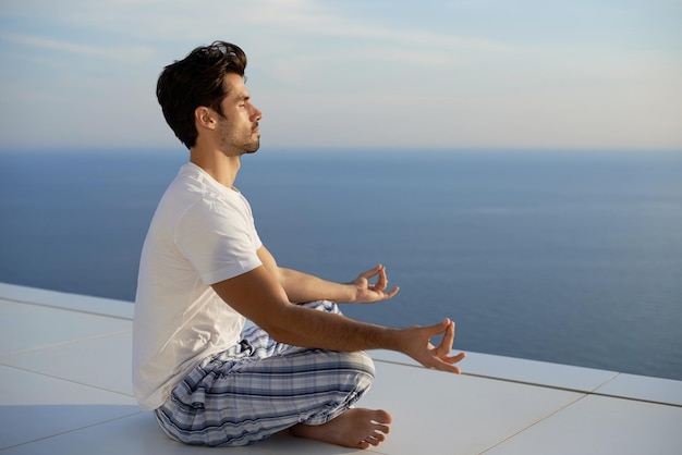 beau jeune homme pratiquant le yoga sur la terrasse de la maison moderne avec l'océan et le coucher du soleil en arrière-plan