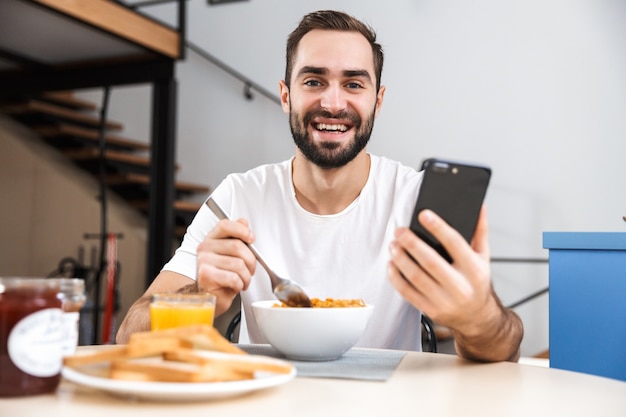 Beau jeune homme prenant son petit déjeuner assis à la cuisine, tenant un téléphone mobile