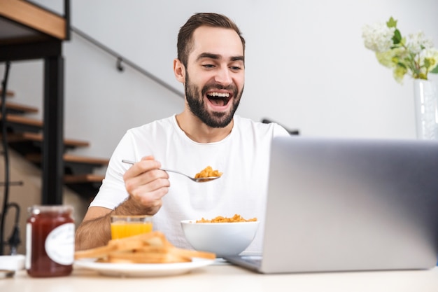 Beau jeune homme prenant son petit déjeuner assis à la cuisine, usinh ordinateur portable