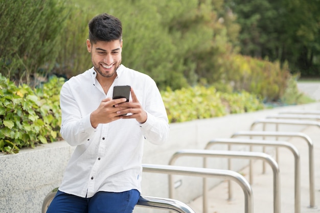 Beau jeune homme de race mixte souriant à l'aide d'un téléphone portable à l'extérieur