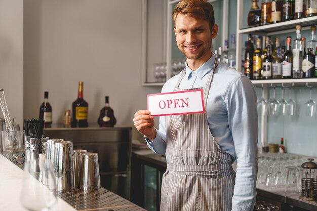 Photo beau jeune homme regardant et souriant tout en démontrant l'enseigne avec le mot ouvert